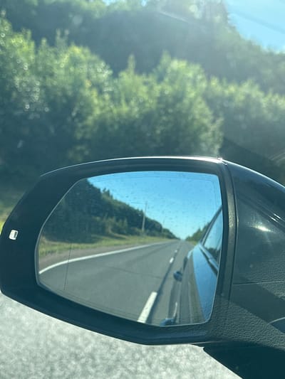 A vehicle's side-view mirror reflects a road with a single lane in each direction, separated by white lines. The road curves to the left, flanked by a grassy shoulder and a dense, green forest on either side. The sky above is a clear blue with some subtle haze. The mirror itself has some water spots or debris on its surface, distorting the reflection slightly. The exterior of the mirror housing is black and textured. No people are visible in the image. The scene appears to be a rural or suburban highway, possibly near Bol'shoe Ondrovo, Russia, given the foliage and road type.