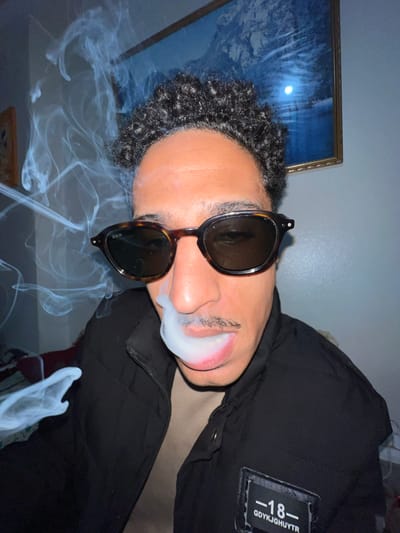 A person with dark curly hair and a mustache is wearing sunglasses and a black jacket. They are exhaling smoke from their mouth. The jacket has a patch on the upper right with the number "-18-" and the text "GDYKJGHUYTR". A framed picture hangs on the wall behind the person. The location is not specified, and it is not possible to determine if it is Tanger طنجة, Morocco.