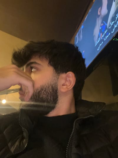 A dimly lit indoor scene captures a man gazing downward with a contemplative expression. His hand partially obscures his face, and a neatly trimmed beard is visible. He's wearing a black jacket. In the background, a television screen displays Arabic script. The lighting is soft, contributing to the subdued mood.