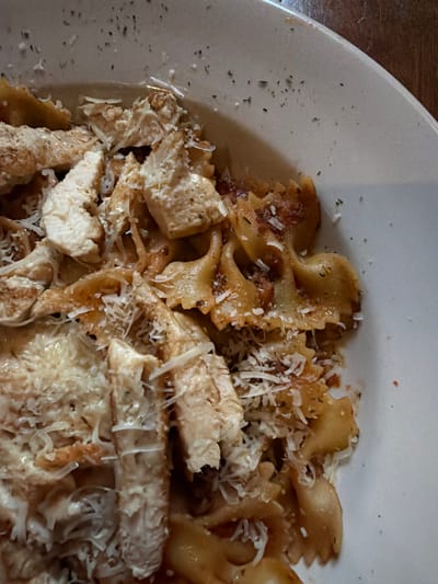A top-down, close-up view shows a partially consumed plate of pasta with chicken and cheese. The pasta is farfalle, commonly known as bow-tie pasta, and appears to be coated in a sauce. Chunks of chicken are scattered throughout the pasta. Shredded cheese is sprinkled on top of the pasta and chicken. The plate is white and has a slightly visible rim on the left side of the frame. The image appears to be taken indoors. The image shows a dish likely served at a restaurant in College Station, United States of America. The photograph shows a pasta dish, specifically featuring farfalle pasta, chicken, and cheese. The ingredients are arranged on a white plate and captured with a close-up perspective. The focus is on the textures and details of the food.