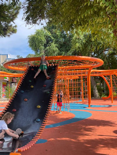 A group of children are playing on an orange and black playground structure in Anapa, Russia. The playground has a climbing wall with colorful grips and a net climbing area. One child is on the climbing wall, wearing a green shirt and shorts. Another child is in the net climbing area, wearing a red shirt and black shorts. A third child is at the bottom of the climbing wall, holding a stuffed animal. The ground of the playground is orange with blue curved lines. Trees with green leaves surround the playground. The sky is blue with some clouds. The lighting suggests it is daytime. The children appear to be enjoying themselves, with smiles visible on some of their faces. The overall impression is a cheerful and active outdoor scene. There is no visible text in the image.
