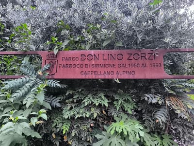 The image shows a rustic, reddish-brown metal sign with cut-out lettering, placed within a lush green natural setting. The sign is mounted on a low barrier or fence, with dense foliage on either side. The text on the sign reads "PARCO DON LINO ZORZI PARROCO DI SIRMIONE DAL 1950 AL 1993 CAPPELLANO ALPINO". This indicates the sign is located in the "Parco Don Lino Zorzi" (Don Lino Zorzi Park) and commemorates Don Lino Zorzi's service as the parish priest of Sirmione from 1950 to 1993, also noting his role as an Alpine chaplain. The surrounding vegetation is abundant and varied, with a mix of leafy plants and some branches covered in what appear to be silvery-green leaves, possibly from an olive tree, and darker, spiky leaves. The overall impression is of a tranquil, natural space within the historic Italian town of Sirmione.