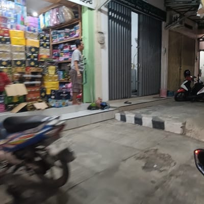 The image shows a street in Kaliwungu, Indonesia, located at coordinates -6.961 latitude and 110.251 longitude. There is a man standing in front of a store, facing a closed gate. The store has shelves stocked with goods and an open doorway to the left of the man. A motorbike is parked in front of the store, facing the closed gate. Behind the motorbike, there is a second, smaller motorbike. The scene is brightly lit, possibly during daylight hours. The street is made of concrete and has a curb and a black and white checkered pattern. The store and the closed gate have a simple facade. The closed gate has a sign with illegible writing. There are boxes and other objects on the ground, possibly debris or discarded items. The man is wearing a light-colored shirt and pants. The motorbikes are both parked without riders. The street is mostly empty with only the man, motorbikes and objects present. There are no signs of any activity or interaction between the man and the motorbikes. The scene is a typical sight in a busy street in a town in Indonesia, possibly in a commercial area.