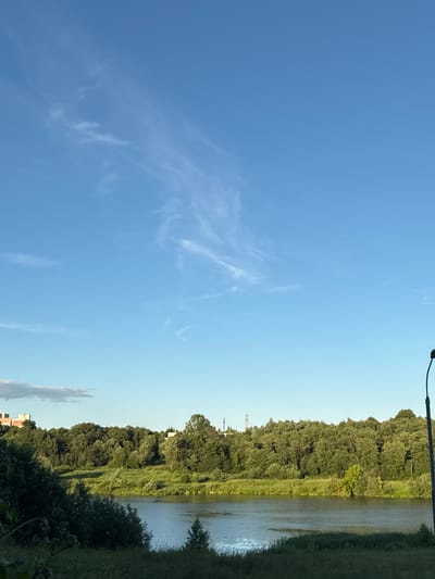 This is a landscape photograph taken in the city of Klin, Russia, during daytime under a clear blue sky with wispy cirrus clouds. The scene depicts a serene natural environment with a body of water in the foreground, possibly a river or a lake, reflecting the light. The water is bordered by lush green vegetation and dense trees forming a natural embankment. On the far bank, a thick line of trees stretches across the horizon, with some buildings and what appear to be communication towers visible in the distance above the tree line. In the right portion of the image, a lamppost is partially visible, suggesting the presence of a park or a developed area adjacent to the natural landscape. The overall atmosphere is peaceful and tranquil, characteristic of a natural setting in Klin.