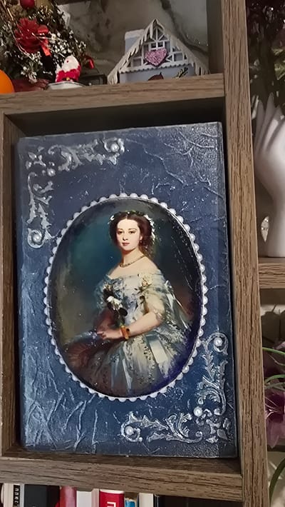 The image shows a decorative frame with a portrait of a woman inside, placed on a shelf. The frame is dark blue with silver embellishments and small white pearls. The portrait is of a woman with brown hair, wearing a white and blue dress and a necklace. She is holding a bouquet of flowers. Above the frame, on the same shelf, are a small Santa Claus figure, a miniature house with a pink heart, and a portion of a decorated Christmas tree. To the right of the frame, a white sculpted hand is visible, and further down, some purple flowers. Below the shelf, books are stacked. The image is taken from a slightly elevated angle, looking down at the shelf. The overall setting appears to be a home decor display. No information about the city of Ақбұлақ, Kazakhstan, can be inferred from the image.