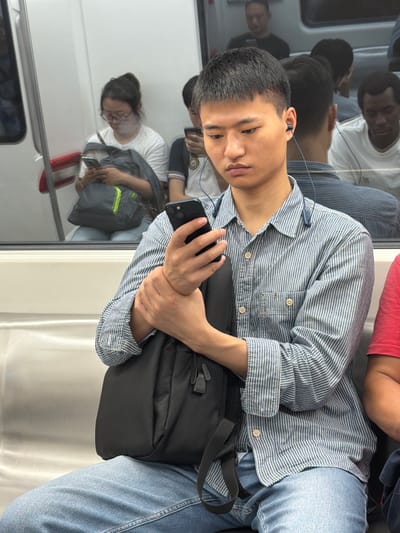 A young Asian man in a blue and white striped shirt and jeans is holding a black backpack and a smartphone. He is wearing earbuds and looking down at his phone. The image appears to be taken inside a public transportation vehicle, likely a train or subway, as suggested by the reflective surfaces and the presence of other passengers visible in the background reflections. The lighting is artificial and the scene is oriented vertically, indicating the photo was likely taken in portrait mode. The man's expression is neutral, and the overall atmosphere seems casual and ordinary. Other passengers are seen in reflections, some also looking at their phones. The scene is indoors and the time of day is not discernible.