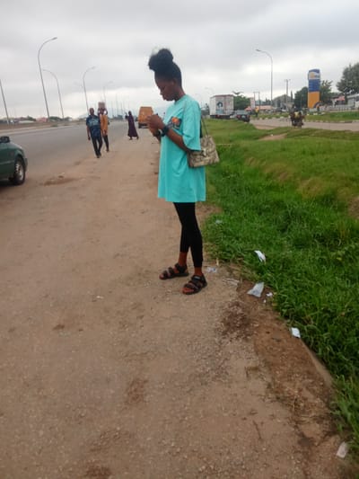 A full shot captures a young Black woman standing on a dirt roadside in Kagini, Nigeria, looking down at her phone. She wears a turquoise t-shirt, black leggings, and black sandals, with a patterned shoulder bag hanging from her right shoulder. Her hair is styled in a bun. Behind her, on the road, a few people walk and carry items, and a yellow van is visible. The environment includes a paved road with vehicles, a grassy verge, and streetlights. The sky is overcast.