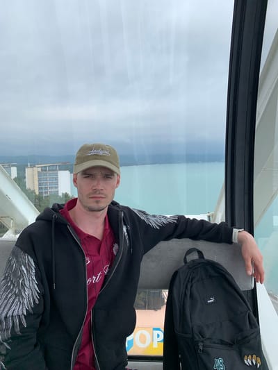 A man is sitting in what appears to be a Ferris wheel gondola. He is wearing a khaki-colored baseball cap with white lettering, a maroon shirt, and a black hooded sweatshirt with silver-colored wing designs on the sleeves. He has a short beard and a serious expression. Next to him is a black backpack. Through the window, a cloudy sky and a body of water are visible, as well as buildings and greenery, suggesting an urban setting near a coastline. Part of a sign with the letters "OP" can be seen through a portion of the window. It’s difficult to determine the precise location in Bichvinta, Abkhazia.