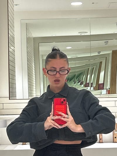 A female subject is depicted taking a mirror selfie in an interior location within Garston, United Kingdom. The subject, positioned centrally and viewed from the upper torso, has dark hair styled in a high bun and wears oval-framed dark glasses. Her mouth is partially open. She is dressed in a dark grey, cropped, collared shirt, with a sliver of abdomen visible, and dark trousers or skirt. Both hands hold a red smartphone, featuring a red textured grip on its rear case, directly in front of her. Multiple rings are visible on her fingers. The background, reflected in the primary mirror, consists of white rectangular tiled walls on the lower section, a white counter surface on the left, and overhead lighting. Behind the subject, a series of mirrors create an infinite reflection effect, showing multiple receding iterations of the interior space, which includes additional tiled walls, light fixtures, and what appear to be wooden partitions or door frames with distant red signage.