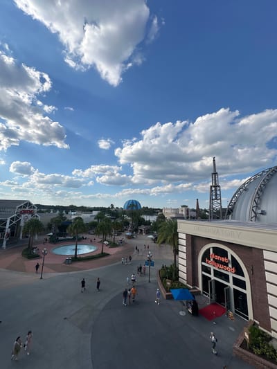 The image captures a scene in Lake Buena Vista, United States, looking over an outdoor promenade, likely in Disney Springs. On the right is the Planet Hollywood Observatory restaurant, a building with a dome-like structure and the "Observatory" lettering visible, along with the Planet Hollywood sign in red. A line of people walks along the walkway, and a fountain is visible in the center. The sky is mostly clear and contains numerous white clouds.