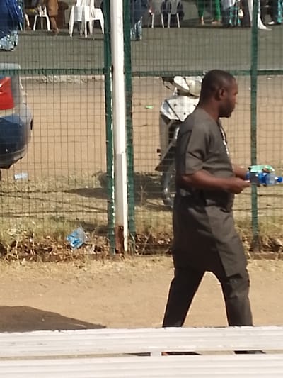 The image depicts an outdoor scene in Sabon Gari East, Nigeria, under clear daylight conditions. An adult male, of medium complexion, is walking from the right to the left side of the frame, positioned in the midground. He wears a dark-colored traditional tunic and trousers, and holds a blue plastic bottle along with another indistinct item in his left hand. Behind him, a green chain-link fence with white vertical support posts spans horizontally across the scene.

Beyond the fence, a blue vehicle's rear section is partially visible on the left, and a white motorcycle or scooter is parked on the right. The ground behind the fence consists of dry, light-brown earth with sparse dry grass and scattered debris, including a crumpled blue plastic bag. In the background, through the fence, multiple white plastic chairs are visible, along with several partially obscured figures, suggesting a distant gathering. A light-colored, slatted structure, possibly a bench or barrier, is present in the immediate foreground, partially visible at the bottom of the image.