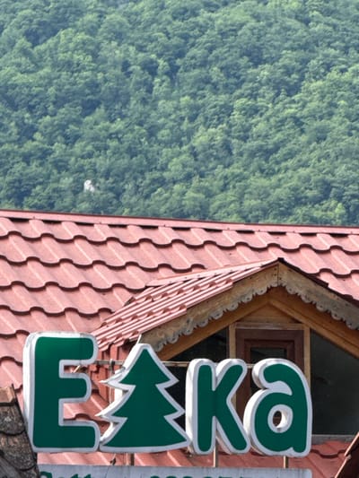 This media file shows a close-up view of a sign with green lettering and a green Christmas tree icon, set against a backdrop of lush green trees and a red, wavy tiled roof. The sign appears to be for a business, possibly a hotel or restaurant, given the stylized lettering and icon. The lettering spells out "eka" and below it is a stylized green Christmas tree and a shape resembling a crown or a stylized building. The letters are three-dimensional, with white outlining.

The scene is outdoors, with the dense green foliage suggesting a natural, possibly mountainous or forested area. The red tiled roof indicates a building, likely situated in a scenic location. The lighting suggests daytime, possibly a bright sunny day given the clear details and contrast. No people are visible in the frame. The overall impression is one of a welcoming establishment in a natural setting. The location context provided is Dilijan, Armenia, a town known for its natural beauty and forests.