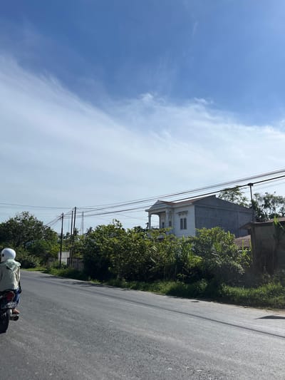 The image depicts a scene from a road in Bukitrata, Indonesia, taken during the daytime under a clear sky with scattered clouds. The perspective is from a moving vehicle, likely a motorcycle, as indicated by the presence of a person on a motorcycle in the lower right corner of the frame. The person is wearing a light-colored shirt and a white helmet.

The road itself is paved and appears to be a two-lane road, with the camera angled towards the right side of the road. Along the right side of the road, there is a dense growth of green foliage and trees, obscuring much of what might be beyond. Further back, a two-story building with a white facade and columns is partially visible, suggesting it's a residential or commercial structure. Electrical wires are strung across the scene, supported by poles, which are common in many areas.

The overall atmosphere is bright and sunny, indicative of a pleasant day. The lush greenery and the style of the building might offer some clues to the specific location within Bukitrata. The license plate on the motorcycle is partially visible, with some text and numbers that are not clearly decipherable. The scene suggests a journey or travel along a rural or suburban road in Indonesia.
