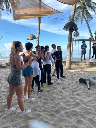 A group of nine individuals, appearing to be young adults, are gathered on a sandy beach, likely in Mũi Né, Vietnam. The scene is outdoors, with palm trees and the ocean visible in the background. The sky is blue with some clouds, and the sun appears to be shining.
In the foreground, a woman with blonde hair tied in a ponytail is facing to the left, her body angled towards a group of other individuals. She is wearing leopard-print athletic shorts and a matching sports bra. To her right, a person with short black hair is wearing a black sports bra and blue athletic shorts. They are holding a dark object, possibly a phone, in their right hand and looking towards the left.
The group continues to the right with several individuals in athletic wear, some in black leggings and tops, and others in white shirts and dark pants. They are all facing generally in the same direction, suggesting they are participating in an activity together, possibly a fitness class or a dance session, given the presence of a disco ball hanging from a palm tree.
Further in the background, two men and a woman are standing on a raised wooden platform. One man wears a blue t-shirt and shorts, while the other wears a white t-shirt and dark pants. The woman on the platform is wearing a dark outfit and white sneakers. There is a swing set structure behind them.
The beach is sandy, with footprints visible. Some logs and stones are present on the sand. Overhead, a light-colored canopy is stretched between poles, providing shade. The overall impression is a casual, active gathering on a tropical beach.