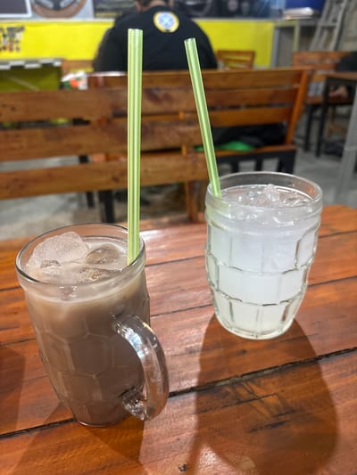 Two beverages with green straws are on a wooden table. The beverage on the left is in a clear mug with a handle and contains a dark brown liquid with ice cubes. The beverage on the right is in a clear glass with a ribbed texture and contains a cloudy white liquid with ice cubes. In the background, a person is seated at a wooden bench. The scene appears to be an outdoor cafe or restaurant.