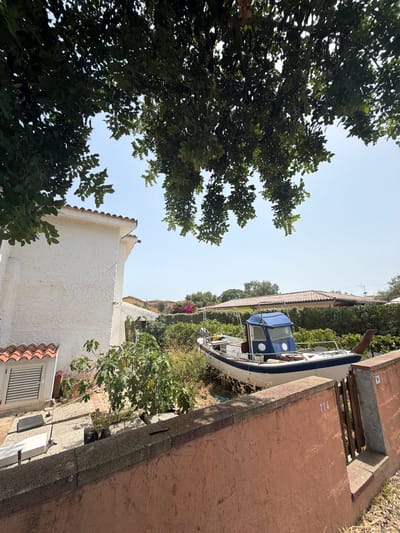 The image shows a view of a boat on land, surrounded by vegetation, with a building and a wall in the foreground. The building, on the left, is white with a textured finish and has a red-tiled roof. A window with horizontal louvers is visible. In the center of the image, a white and blue motorboat is parked on what appears to be a gravelly or overgrown area. The boat has a cabin structure with blue panels and a white roof. Behind the boat, there are dense green bushes and trees, and in the distance, a house with a red-tiled roof is visible. A peach-colored wall runs along the right and bottom edges of the image, with a section of a wooden gate visible. The number "114" is painted in white on the wall near the gate. Above the scene, lush green tree branches with leaves hang down, partially obscuring the sky, which is clear and blue. The scene is lit by bright daylight, suggesting it is daytime. The overall impression is of a semi-rural or coastal setting, possibly a private property where the boat is stored. Santu Diadòru/San Teodoro is a popular tourist destination on the coast of Sardinia, Italy, known for its beaches and natural beauty, which could be consistent with this type of scenery.