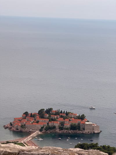 This is a high-angle, vertical, medium shot of Sveti Stefan, a picturesque islet and fortified village in Montenegro, connected to the coast by a narrow causeway. The islet is densely packed with stone buildings topped with red tile roofs, nestled amongst lush green trees. Several small boats and a larger white yacht are visible in the calm, blue-gray water surrounding the island. The causeway, a sandy beach, and a walkway are populated with many small figures of people, suggesting a pleasant, bustling day. The scene is bathed in natural light, implying it's daytime, and the sky appears overcast, contributing to the muted tones of the water and the soft lighting. The foreground features the rough, textured surface of a rocky cliff, adding a sense of depth and perspective to the view. The overall mood is tranquil and scenic, capturing a popular tourist destination. There is no visible text in the image.
