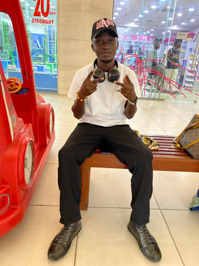 A young Black male wearing a black baseball cap, a white t-shirt, black pants, and black sneakers is sitting on a wooden bench in what appears to be a shopping mall in Ikeja, Nigeria. He has headphones around his neck and is holding up two fingers on his right hand and pointing two fingers on his left hand in a peace sign. A red toy car is to his left, and a patterned handbag rests on the bench to his right. Behind him, through a glass storefront, a pharmacy is visible with shelves stocked with products and several other shoppers. A sign in the background indicates "20% DISCOUNT." The floor is tiled in a light color.