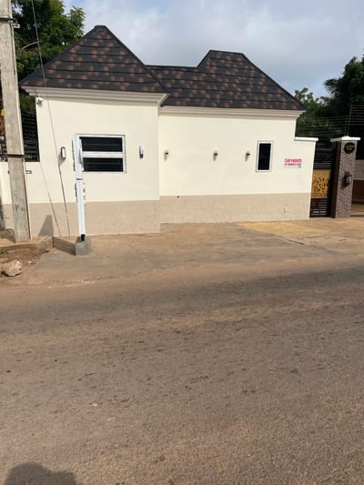 The image shows the exterior of a modern house in Narayi, Nigeria. The house is painted cream and has a dark brown tiled roof. There are two windows visible on the front of the house, both with security bars. A sign that reads "CAR PARKED AT OWNER'S RISK" is posted on the wall near a gated entrance. The gate is made of metal with an ornate design and is flanked by brick pillars. A street light is visible on one of the pillars. In front of the house is a paved driveway and a section of road. Trees and other vegetation are visible in the background. The sky is overcast.