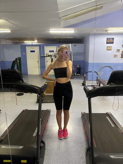 The image depicts a young woman with blonde hair, wearing a black crop top and black biker shorts, standing in a gym. She is holding a pink smartphone and taking a selfie in a large mirror. The gym appears to be in a modest facility, with two treadmills visible in the foreground. In the background, there are gym equipment, weights, and posters on the wall. The overall impression is of a functional, somewhat dated gym environment. The specific location in Ust-Ilimsk, Russia, cannot be determined from the image alone.