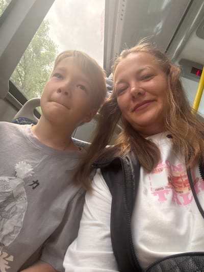 A woman and a young boy are posing for a selfie. The woman is wearing a white T-shirt and a black jacket. The boy is wearing a grey T-shirt with a print. They appear to be inside a bus. The location may be the city of Dmitrov, Russia.