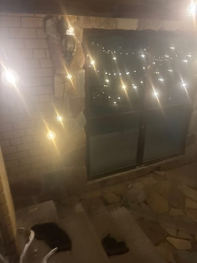 The image captures an outdoor scene at night, illuminated by string lights and a traditional outdoor lantern. The environment appears to be a patio or entryway with a stone and brick facade. A large window is visible, reflecting the string lights, which are also draped across the scene, creating a festive or ambient atmosphere. In the foreground, on a stepped surface, a dark-colored object resembling a blanket or piece of fabric is present, alongside what appears to be a shoe. Another object, possibly a white bag or cloth, is partially visible next to the dark object. The ground is paved with irregular stone tiles. The scene is set in the city of Orekhovka, Russia.