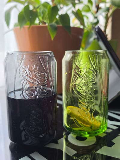 This media file displays two glass objects, each shaped like a Coca-Cola can. One glass is a vibrant lime green and appears to contain a yellow substance, possibly lemon slices. The other glass is split between clear and black, with the Coca-Cola logo embossed on both. The objects are placed on a white reflective surface. In the background, blurred green foliage from a plant is visible, along with a brown terracotta pot. The overall lighting suggests daytime. The objects are positioned horizontally, creating a reflection on the surface below. The Coca-Cola logo is prominently visible in raised lettering on both glasses, with a distinct cursive font. The context provided indicates the location is St. Gallen, Switzerland. No people are visible in the image.