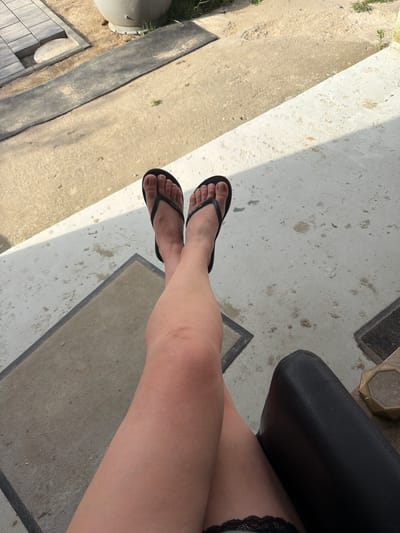 This is a close-up, overhead shot of a person's legs and feet, wearing black flip-flops. The person's legs are crossed and resting on a concrete surface, with a black chair visible to the right. The background shows a patterned paved area with some scattered debris and a large grey planter. The overall lighting suggests an outdoor setting, possibly a patio or balcony, during daylight hours. The image does not contain any elements that would allow for its specific location to be identified as Szczecin, Poland.