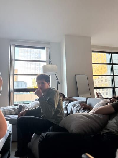 The image depicts a modern, brightly lit apartment interior in a dense urban area, identified as being in Boston, United States. A young adult male with dark curly hair, wearing an olive green long-sleeved shirt and dark pants, sits upright on a grey sectional sofa in the left-center foreground. His right hand is raised to his mouth, possibly holding a small, dark object. He is looking directly towards the camera. To his right, on the same sofa, a second individual is partially obscured, lying down and covered by a light-colored blanket or pillow. Further to the right, a third individual is reclined on the sofa, mostly covered by a grey pillow, resting or sleeping. A fourth person's arm and shoulder are partially visible on the far left of the frame.
The background features large windows revealing multiple high-rise city buildings under bright daylight, including one with a distinct yellowish facade. A modern floor lamp with a white drum shade stands behind the central seated figure. A dark-framed rectangular mirror or artwork leans against a white wall to the right of the lamp. The apartment walls are white, and the visible flooring is dark. The overall scene suggests a casual, relaxed daytime setting.