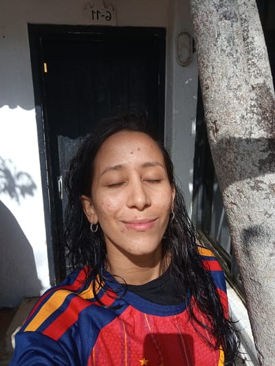 This is a portrait of a young woman with her eyes closed, bathed in bright sunlight. She has long, dark, wet hair and her skin shows signs of acne. She is wearing a red, blue, and yellow striped athletic shirt. The background features a dark door, a white wall with a subtle shadow of foliage, and a large, textured tree trunk. A small, oval light fixture is visible on the wall. The overall impression is of a casual outdoor moment, likely in a warm climate, given the sunlight and the presence of the tree. The time of day appears to be daytime, possibly late morning or early afternoon, due to the strong shadows. The woman's closed eyes and slight smile suggest a feeling of peace or enjoyment of the moment. A small sign above the door displays what appears to be Arabic numerals or letters. The location context provided is Porlamar, Venezuela, which aligns with the sunny and potentially tropical setting.