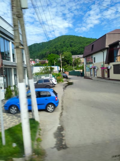 The image captures a street scene in Agoy, Russia, on a partly cloudy day. A bright blue hatchback car is parked on the left side of the asphalt road. Further down the road, a dark-colored SUV and a white SUV are also visible. A modern building with a brown roof and cream-colored walls stands on the right side of the road, featuring windows and signs. In the background, a lush, green forested hill dominates the landscape under a bright blue sky dotted with white clouds. A tall utility pole with wires runs vertically on the left side of the image. The overall atmosphere appears casual, with no specific event or activity prominently featured, beyond the presence of parked vehicles and a couple of indistinct figures walking further down the street. The motion blur suggests the image was taken from a moving vehicle.