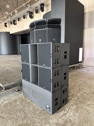 In Baku City, Azerbaijan, a stack of professional audio speakers is positioned on a textured carpeted floor. The speakers are arranged in a vertical configuration, with larger units at the bottom and smaller units on top. The speakers are dark gray and appear to be part of a sound system for an event or performance space. In the background, a large, dark structure and cylindrical columns are visible, suggesting an indoor venue. Overhead, a grid of lighting fixtures hangs from the ceiling, some with clear glass globes and others with dark casings, likely spotlights. The overall environment appears to be a contemporary exhibition hall, concert venue, or convention center.