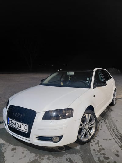A white Audi A3 hatchback is parked on a wet concrete surface at night in Borovan, Bulgaria. The car's headlights are on, illuminating the front of the vehicle and the ground around it. The license plate reads "CB4633TC". The background is dark, with the silhouette of a leafless tree visible. There are no people present in the image. The scene appears to be an outdoor parking area, possibly a gas station or a service area, given the wet ground and the lighting.