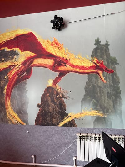 The image depicts a wall mural of a red dragon engulfed in flames, with mountainous terrain in the background. A black, multi-faceted object, possibly a motion capture sensor, is mounted on the wall above and to the left of the dragon mural. Below the mural, a section of a grey textured wall is visible, featuring white piping and a white radiator. A black computer monitor is positioned on the right side of the frame, partially obscuring the radiator and the wall. The overall scene appears to be an indoor space, likely a room, with the mural serving as a prominent decorative element. The location of Chuguyevka, Russia is not evident from the visual information in the image.