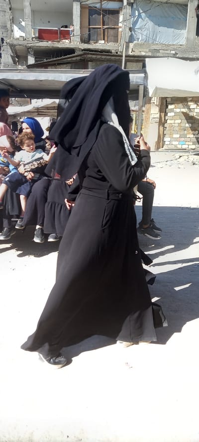 The image shows a group of people, including adults and children, in an outdoor setting that appears to be a war-torn or damaged urban area. The subjects are partially obscured by shadows and the perspective of the camera, which is tilted. Several people are wearing black clothing, including long dresses or robes, and some are wearing headscarves. One person in the foreground is wearing a blue headscarf and a patterned garment. Children in the lower part of the frame are also visible.
The background suggests a damaged environment, with visible concrete structures, exposed rebar, and rubble. The buildings in the background appear unfinished or destroyed, with some covered by white sheeting. The scene is lit by bright sunlight, casting long shadows on the ground, indicating it is daytime. The overall atmosphere seems somber, possibly due to the surrounding destruction.
The image is taken in Khan Yunis, Palestinian Territories. No specific activity or event is clearly discernible beyond people gathering or moving through the area. There is no visible text in the image.