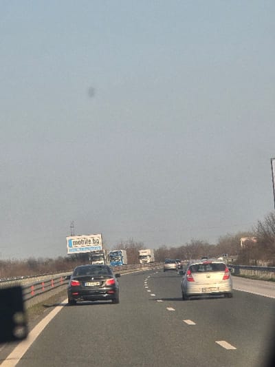 The image depicts a multi-lane highway scene under a clear, light blue sky, located in the vicinity of Trilistnik, Plovdiv Province, Bulgaria. The perspective is from a vehicle on the highway, looking forward.
In the immediate left travel lane, a dark-colored BMW E60 5 Series sedan, bearing Bulgarian license plate 'PP 4842 HM', is positioned near the solid white line separating the lane from the paved shoulder. A metal guardrail with orange delineator posts runs parallel to the road on the left. In the right travel lane, further behind the BMW, a silver Hyundai i30 hatchback with Bulgarian license plate 'PB 2074 KA' is visible.
The background shows additional vehicular traffic, including several white and blue heavy goods vehicles and passenger cars, receding into the distance. A large billboard displaying 'mobile.bg' in blue text on a white background is situated on the left side of the highway, above the guardrail. The roadside environment features bare deciduous trees and shrubs beyond the guardrails.
No individuals are visible. A small, dark particulate spot is present in the upper central sky region of the frame. A blurry, dark rectangular object is partially visible in the lower left foreground, consistent with an interior vehicle component.