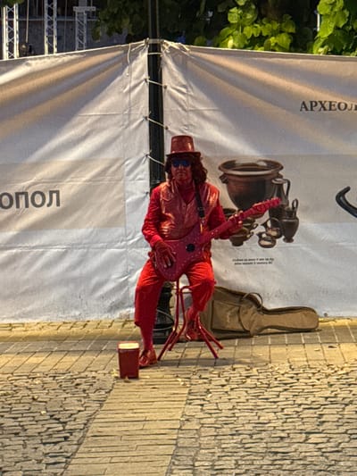 This media file shows a performer dressed in a vibrant red costume, sitting on a chair and playing a red electric guitar. The performer wears a hat and sunglasses, adding to their theatrical appearance. A guitar case, likely for the instrument being played or another, is visible leaning against a textured stone wall. The background features a large white banner with text and graphics, possibly an advertisement or informational display for an event or location. The scene appears to be outdoors, with cobblestone paving visible on the right side, indicating a street or plaza. The lighting suggests it is daytime, with sunlight casting shadows. The overall impression is of a street performance or a festival event, potentially in a historic or tourist area. The word "АРХЕОЛ" is visible in large letters on the banner, and below it "ОПОЛ", suggesting a connection to archaeology or an archaeological site. Given the location context of Sozopol, Bulgaria, it is likely this scene is part of a cultural event or festival in that ancient town.