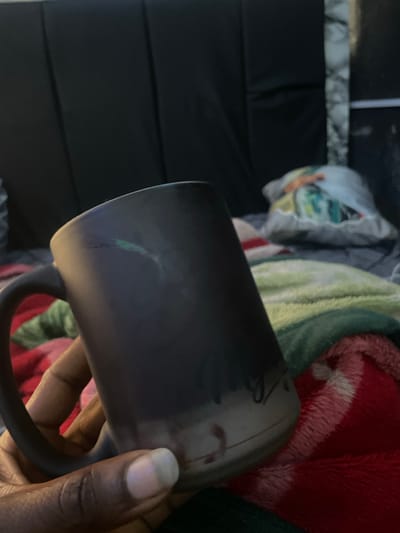 A dark-colored mug is held by a person's left hand, with their thumb resting on the mug's rim and fingers curved around the handle. The mug is dark, with some lighter markings and a slight gradient towards the bottom. In the background, a dark, padded headboard is visible, partially obscured by a patterned throw pillow. Below the headboard, several blankets are spread out, including one with green and yellow stripes and another with red and white stripes. The scene suggests an indoor setting, possibly a bedroom or living room. The specific location within New Stadium, Nigeria, cannot be determined from the image alone.