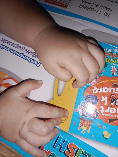 The image displays a close-up, overhead view of a child's hands interacting with educational materials, set in the city of Alue Mudem, Indonesia. Two small, light-skinned hands are visible. The left hand (viewer's left) has its index finger resting on a yellow, contoured object featuring a perforated speaker grille. This yellow object is partially covering a blue-colored book or box. The right hand (viewer's right) is positioned with its fingers resting on the same blue book/box and the yellow object.
The blue book/box features colorful illustrations including cartoon animals (e.g., a cow, a cat, a mouse), a wooden chair, musical notes, and a green fruit (possibly an avocado). Visible text on this item includes "Belajar SUARA" (Learn Sound), "Indonesia English Mandarin" indicating language options, and "Pelajaran yang menarik" (interesting lessons). Further down, partially visible text on another blue item reads "ENGLISH". In the background, on what appears to be a white box or surface, manufacturing details "NO. TY-000682" and "MADE IN CHINA" are visible, along with an upside-down "PERINGATAN!" (Warning!). The scene suggests an indoor environment, likely a tabletop where a child is engaged with interactive learning toys.