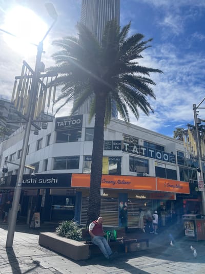 In Surfers Paradise, Australia, a sun-drenched street scene unfolds, dominated by a tall palm tree casting a shadow across the sidewalk. Above, a modern building displays prominent signage for "SKIN FX TATTOO Since 1985" and "4siteprotection.com.au." To the left, another sign reads "sushi sushi." Below, an orange awning stretches across the storefronts of "Creamy Italiano," an ice cream parlor. Several individuals are present: a man in a red vest and blue jeans sits on a bench, seemingly engrossed in his phone, while other people walk by or stand in small groups near the establishments. Pigeons peck at the ground in the foreground. The environment is urban, with paved sidewalks, streetlights, and other buildings visible in the background, under a bright blue sky with scattered clouds.