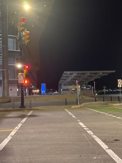 This is a nighttime exterior photograph of a street in Vancouver, Canada, specifically near the Vancouver Convention Centre at Canada Place.

In the foreground, a street intersection is visible with clear lane markings, including solid and dashed white lines. A green painted area, likely a bike lane, is present on the right side of the image. The pavement is asphalt.

To the left, a traffic light pole is prominently featured. The traffic signal for vehicles is showing red, and the pedestrian signal is displaying a red hand, indicating "Don't Walk." Below the traffic lights is a sign that reads "VEHICLES YIELD TO BICYCLES." A lamppost with a bright light illuminates the area. Behind the traffic light pole, a modern building with a glass facade and a patterned ceiling can be seen.

In the midground, a wide set of stairs leads up towards a covered outdoor area. This area appears to be an entrance or waiting zone, with a modern, sloped roof structure supported by pillars. A sign with the text "VANCOUVER CONVENTION CENTRE" is visible on a wall behind the stairs. Further in the background, across a body of water, a cityscape with illuminated buildings is visible, suggesting a waterfront location. The sky is dark and clear.

The scene is devoid of people or any ongoing activity, presenting a static, urban environment at night. The lighting is a mix of artificial sources from streetlights, building interiors, and the traffic signals. The overall impression is one of a quiet, well-lit urban space.