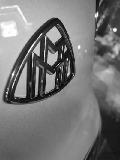 The black and white media file shows a close-up, angled shot of a Maybach emblem on a car. The emblem is metallic and features the intertwined "M" logo. It is set against the car's body, which appears to be a light color, possibly white or silver, and has a smooth, reflective surface. The background is out of focus, showing blurred shapes and lights, suggesting an outdoor setting at night or in low light conditions, possibly in a parking lot or on a street. Some of these blurred lights resemble reflections or streetlights. The overall impression is one of luxury and automotive detail. No people are visible in the frame. The image does not contain any discernible text.
