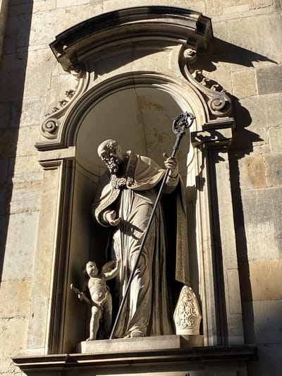 The image shows an exterior sculpture of Bishop Benno, located on the wall of the Katholische Hofkirche in Dresden, Germany. The sculpture is rendered in stone and depicts the bishop lying in repose, holding a crozier. An angel figure is positioned at the base of the bishop's likeness. The sculpture is enclosed in a rounded alcove with elaborate ornamentation. The scene is lit by daylight, creating shadows on the wall and sculpture.