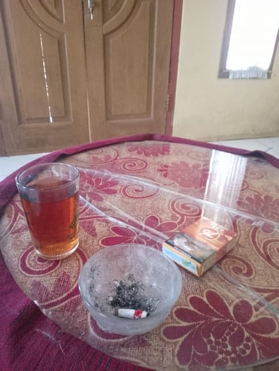 This image captures a still life scene on a circular table with a patterned cloth and a glass top. On the table, there is a glass of amber-colored liquid, likely tea, with a faint green design on its side. Adjacent to the glass is an ashtray containing cigarette ashes and a single cigarette butt. To the right of the ashtray and glass, a pack of cigarettes labeled "76 APEL" is positioned. The background shows a wooden door with vertical paneling and a small window with a light-colored curtain. The lighting suggests an indoor setting, possibly a home or a simple establishment. The overall impression is of a moment of pause, perhaps a break for a cigarette and tea. The presence of cigarettes and an ashtray suggests a smoking area, common in many Indonesian households and public spaces. Given the context of Sukamanah, Indonesia, this scene could be representative of a casual, everyday setting.