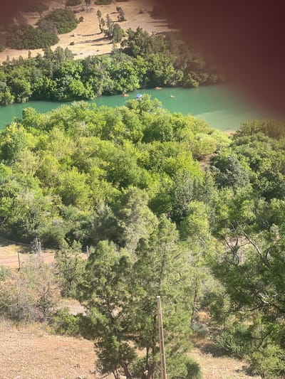 The image shows an aerial view of a river surrounded by dense green vegetation, likely in a forested area. The river is a light green color, and there appear to be a few small boats or kayaks on the water. On the other side of the river there is a grassy terrain. There are trees and shrubbery between the river and the grassy area. At the top of the image, there is a patch of discoloration, possibly caused by a light leak or lens flare. It is impossible to ascertain from the image if this is the specific location of Bin El Ouidane, Morocco.