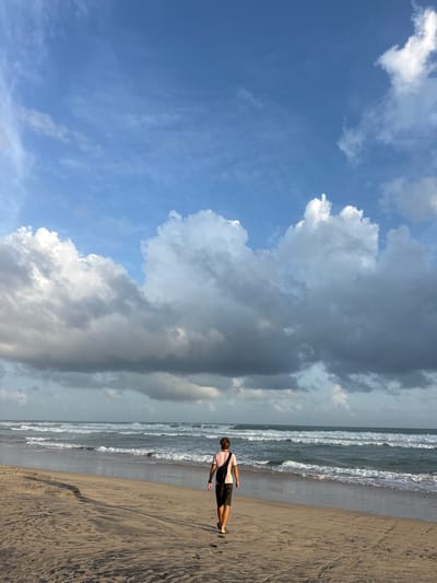 The image depicts a lone individual walking on a sandy beach, facing the ocean. The person is wearing a light-colored t-shirt and dark shorts, with a dark bag slung across their chest. They are positioned in the lower right portion of the frame, facing left, with their back mostly to the viewer.
The scene is set on a beach with a sandy shore that shows footprints and ripples from receding waves. The ocean is visible with white-capped waves rolling in. The sky is a prominent feature, with a mix of bright blue sky and large, dramatic clouds. Some clouds are white and puffy, suggesting fair weather, while others are darker and more ominous, hinting at potential rain or a change in weather.
The lighting suggests it is either early morning or late afternoon, as the sun is not directly overhead. The overall mood is peaceful and solitary, with the vastness of the ocean and sky surrounding the single figure. There is no visible text in the image. The location context provided is Kathaluwa, Sri Lanka, which aligns with a tropical beach setting.