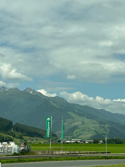 The image shows a landscape in Bruck an der Großglocknerstraße, Austria. In the foreground, a highway runs horizontally, bordered by a grassy median with a guardrail. Beyond the highway, a vast green field stretches towards a collection of industrial buildings on the left and a cluster of houses and farm buildings on the right. Two green flags with white lettering are prominently displayed in the middle ground. The flags appear to be for a business named "LAGERHAUS". In the background, a majestic mountain range dominates the scene, with lush green forests covering its lower slopes and snow-capped peaks reaching towards a sky filled with scattered white clouds. The overall scene conveys a sense of rural tranquility and the majestic beauty of the Austrian Alps.