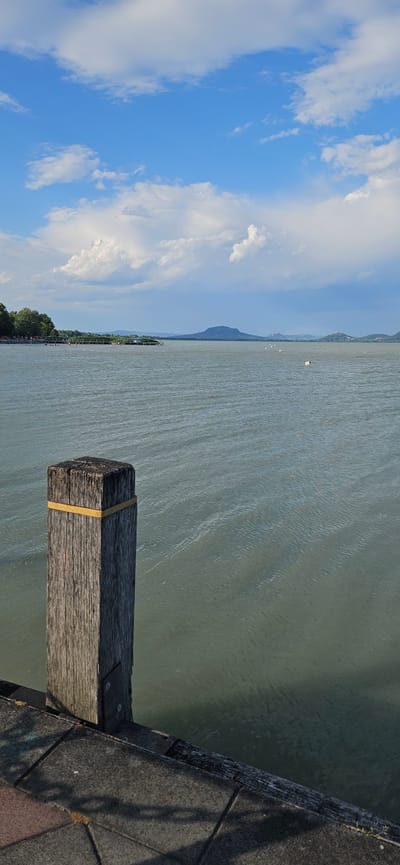 The image depicts a calm lake scene with a wooden bollard in the foreground. The bollard, made of weathered grey wood, has a yellow band around its top and is bolted to a stone pathway. The pathway, constructed of dark grey and reddish paving stones, leads to the edge of the lake. The lake water is a muted green, with gentle ripples creating a textured surface. In the mid-ground, a thin wooden pier or walkway extends into the water, lined with reeds and vegetation on the left bank. Beyond the pier, distant land formations are visible, including trees on a far shore and a prominent, cone-shaped mountain in the distance. The sky above is a vibrant blue, dotted with fluffy white clouds. The overall scene suggests a peaceful daytime environment. This location is identifiable as the Balaton shore in Balatongyörök, Hungary, a popular tourist destination known for its scenic views of Lake Balaton and the surrounding landscape, including the Tapolca Basin.