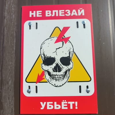 The image displays a warning sign featuring a skull and crossbones motif. The sign is predominantly red and white, with a yellow triangle behind the skull. Red lightning bolts are depicted, one above and one below the skull. The text on the sign reads "НЕ ВЛЕЗАЙ!" (Do not enter!) at the top and "УБЬЁТ!" (Will kill!) at the bottom. The sign is affixed to a brown, corrugated surface, possibly a metal fence or wall. The location in the city of Sirius, Russia, is not discernible from the image itself.