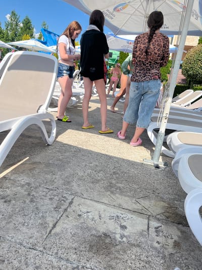 Three young women stand near lounge chairs in an outdoor area, possibly a water park or resort, under a large umbrella. The woman on the left has long brown hair and wears a white t-shirt and denim shorts. The woman in the middle has dark hair and wears a black top and black shorts. The woman on the right has dark hair in a braid and wears a leopard-print top and denim capris. She also wears pink Crocs. The ground is paved with concrete slabs. In the background, there are lounge chairs, trees, and a blue waterslide. The sun is shining brightly. The scene is likely located in Ravda, Bulgaria, a popular resort town on the Black Sea coast known for its beaches and water parks.